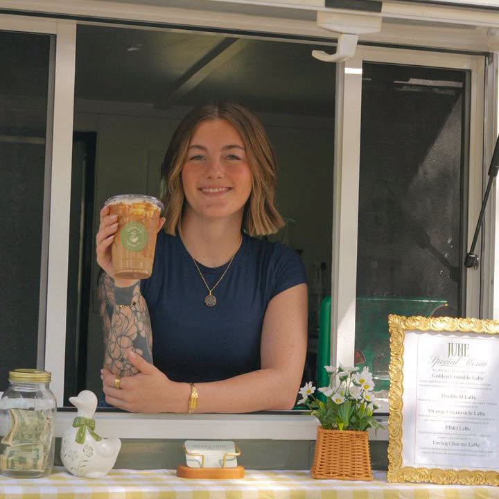 The owner of Double H Coffee Co. smiling at the trailer's service window, holding an iced latte above a gingham tablecloth with fresh flowers